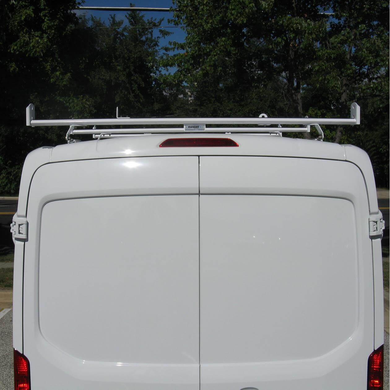 The back of a white van with closed doors, featuring a roof rack against a background of trees and clear sky.